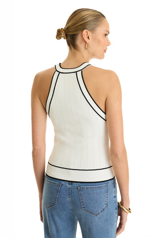 A woman with blonde hair in a sleek bun models the Martha Button Halter Tank in white—a sleeveless, ribbed knit top with black trim—paired with blue jeans. She faces away, showcasing the back details against a white background.