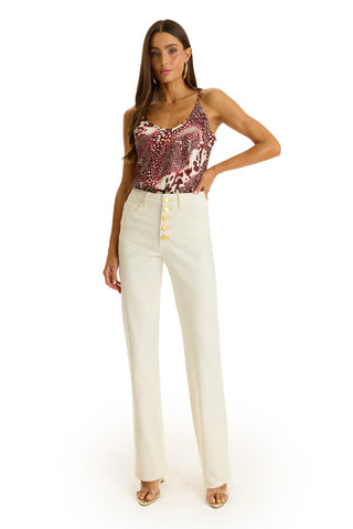 A woman poses against a white background, wearing a red and white patterned sleeveless top, the Aubrey Bootcut Jean in ivory with large buttons, and open-toe heels.
