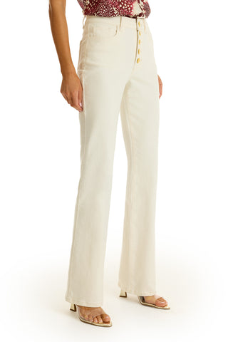 A woman’s lower body is shown against a white background, styled in the Aubrey Bootcut Jean - Ivory, featuring a high waist and gold buttons, paired with a printed blouse and open-toed heeled sandals.