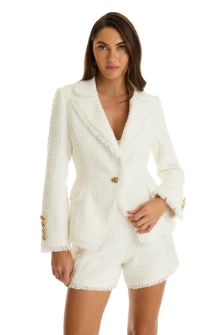 A woman poses confidently against a white background, wearing the Giselle Single Breasted Tweed Blazer in white, featuring padded shoulders, gold buttons, frayed edges, and matching high-waisted shorts. One hand rests in her pocket.