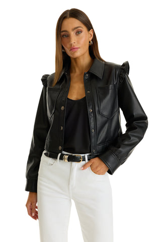 A woman with straight brown hair wears the Chantal Ruffle Sleeve Leather Jacket in black over a black top and white high-waisted jeans, posing with one hand in her pocket against a white background.