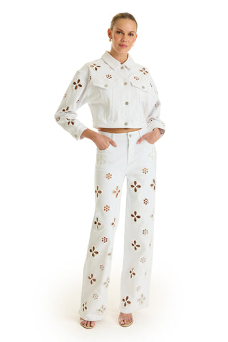 A woman with her hair pulled back stands with hands in pockets, wearing the Kaia Pearl Jean Jacket in white, high-waisted jeans with brown and beige floral cutouts, and open-toed heels.