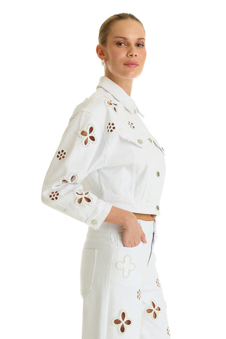 Woman wearing the Kaia Pearl Jean Jacket in white, featuring floral cut-out details, posing with one hand in her pocket against a white background.