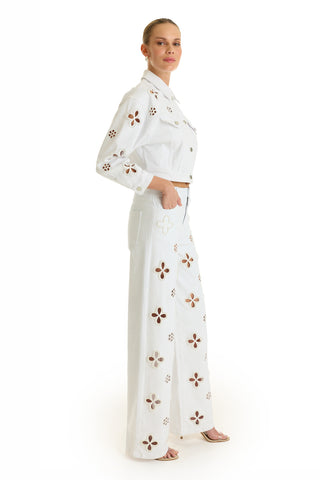 A woman in a side pose wears the Kaia Pearl High Rise Jean in white, featuring cut-out flower patterns and pearl embroidery, paired with a white jacket and heels against a white background.