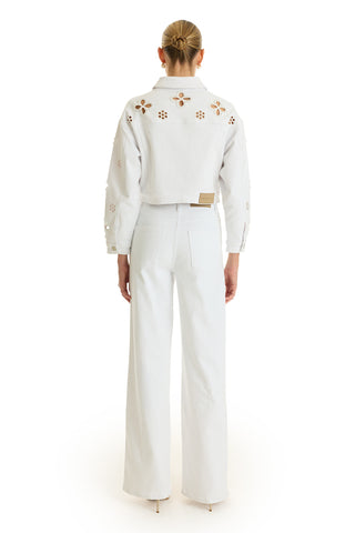 A person wearing the Kaia Pearl High Rise Jean - White stands with their back to the camera, paired with a cropped white denim jacket featuring floral cutouts. Their hair is tied in a neat bun against a plain white background.