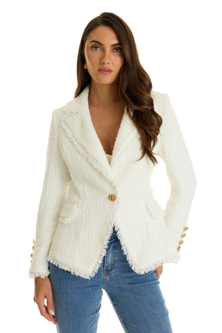 A woman with long brown hair wears the Giselle Single Breasted Tweed Blazer in white, featuring gold buttons and padded shoulders. She pairs it with a white top and blue jeans, standing confidently facing the camera.