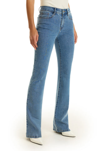 A person wears the Aria Flare Jean in medium blue with white pointed-toe shoes, standing against a white background. The upper body and face are not shown.