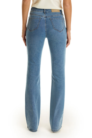 A person is shown from the back wearing the Aria Flare Jean in medium blue, featuring two back pockets and a leather patch on the waistband, paired with a cream-colored top.