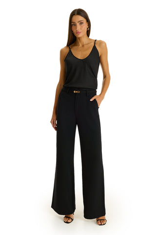 A woman stands against a white background wearing the Martina Belted Trouser in black, paired with a black tank top, heeled sandals, and hoop earrings. One hand is tucked in her pocket.