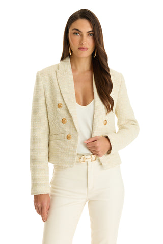 A woman stands against a white background, wearing the Gabrielle Tweed Double Breasted Blazer in beige with gold buttons over a white top, paired with cream pants and a belted waist.