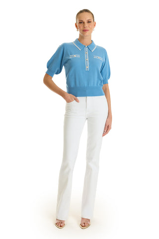 A woman poses against a white background wearing the Bobbi Polo Shirt in Serene Blue with white trim, styled with white high-waisted straight-leg pants and open-toe heels. Her hair is tied back for an elevated casual look.