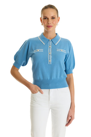 A woman wears the Bobbi Polo Shirt in Serene Blue—featuring short sleeves, white trim, buttons, and two chest pockets—paired with high-waisted white pants. She stands against a white background, smiling softly.