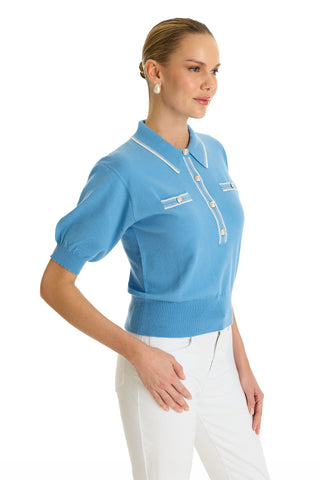 A woman smiles in the Bobbi Polo Shirt - Serene Blue, featuring a collar and decorative buttons, paired with white pants. She poses with one hand on her hip against a white background—an elevated take on casual wear.