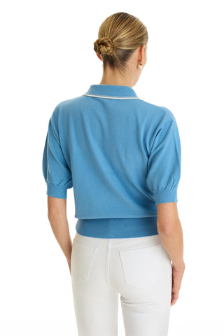 A woman with blonde hair in a low bun is seen from behind, wearing the Bobbi Polo Shirt in Serene Blue and high-waisted white pants, creating an elevated casual look against a white background.