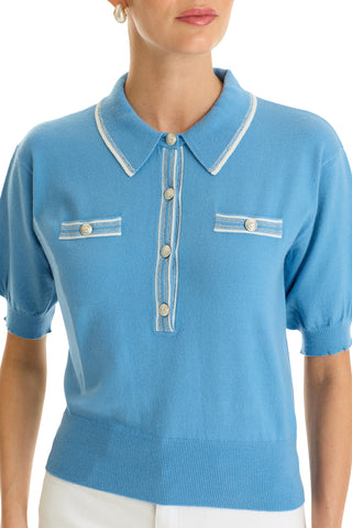 A woman wears the Bobbi Polo Shirt in Serene Blue, a chic knit top with a collar, decorative buttons, white trim, and two faux chest pockets. She pairs it with white pants; only her torso and part of her face are visible.