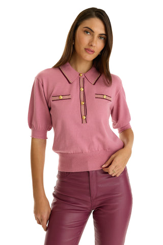 A woman in a Dusty Rose Bobbi Polo Shirt featuring gold buttons and matching high-waisted pants stands against a white background, facing forward with one hand on her hip—an elevated casual wear look.