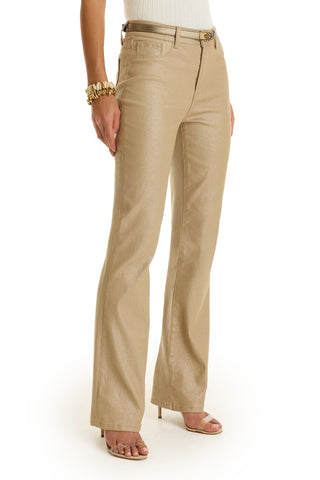A person wears the Aria Coated Flare Jean - Champagne, featuring a high-rise flare fit, styled with a belt, white sleeveless top, gold bangles, and beige open-toe heels, standing against a white background.