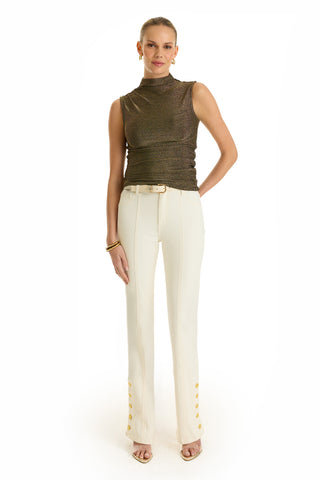 A woman stands against a white background, wearing the Mia Shimmer Tank in gold, high-waisted white pants with gold buttons at the ankles, beige heels, and gold hoop earrings.