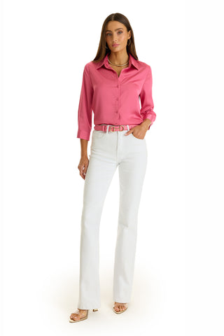 A woman with long brown hair and a neutral expression stands against a white background, wearing the Sara Silk Blouse in Petal Pink with a high-low hem and three-quarter sleeves, paired with white high-waisted pants and nude heeled sandals.