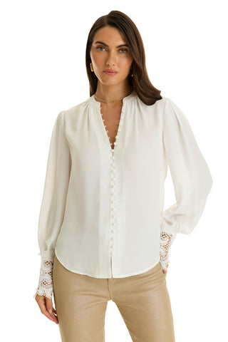 A woman with long brown hair wears the Blake Lace Sleeve Blouse in white, featuring lace sleeves, covered buttons, and lace cuffs. She pairs it with light beige pants and stands facing forward, one hand by her side.