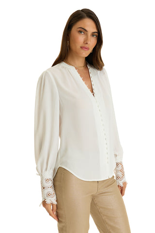 A woman models the Blake Lace Sleeve Blouse in white, featuring decorative buttons and lace cuffs, paired with light beige pants, against a white background.