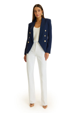 A woman faces forward against a white background, wearing the Sasha Double Breasted Blazer in navy with gold buttons, paired with a white top, white high-waisted pants, and beige open-toe heels.