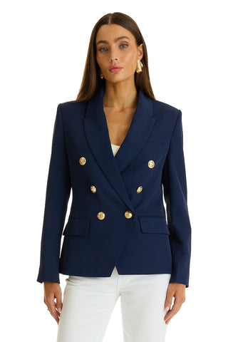 A woman stands against a plain white background wearing the Sasha Double Breasted Blazer in navy with gold buttons over a white top and white pants.
