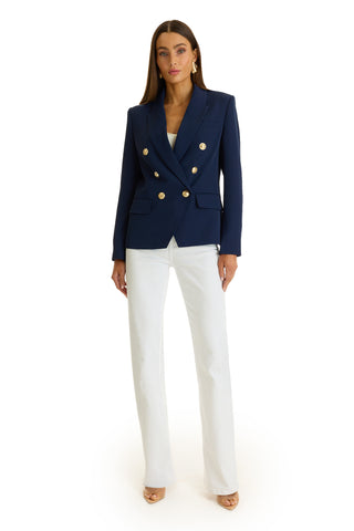 A woman poses against a white background wearing the Sasha Double Breasted Blazer in navy, styled with white pants, nude heels, and hoop earrings.