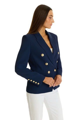 A woman wears the Sasha Double Breasted Blazer in navy with gold buttons and white pants, standing sideways against a white background.