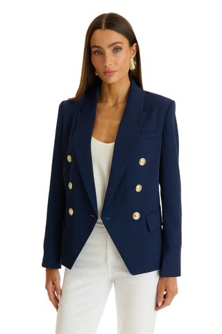 A woman wears the Sasha Double Breasted Blazer in navy with gold buttons over a white top and white pants, standing against a plain white background.