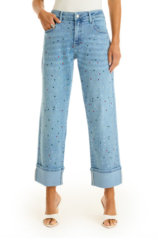 Wearing the Ren Cropped Cuff Jean - Confetti, featuring light blue denim with paint splatters and a rainbow gem finish, paired with white heeled sandals, a person stands against a white background.