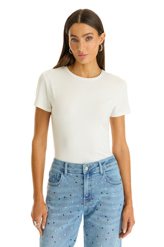 A woman with long brown hair wears the Amaya Ultra-Soft T-Shirt in white, tucked into light blue jeans with small colorful dots, standing against a white background.