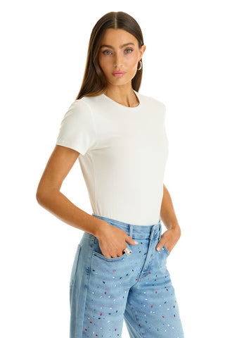 A woman with long brown hair wears the Amaya Ultra-Soft T-Shirt in white and light blue jeans with small colorful dots, standing with her hands in her pockets against a white background.