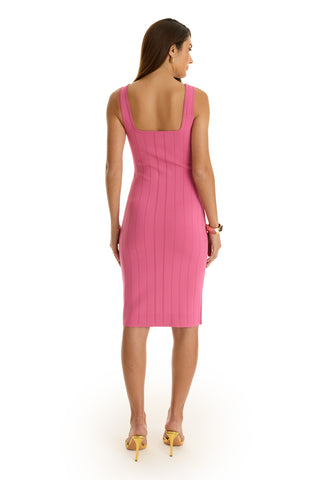 A woman with long brown hair faces away, wearing the form-fitting Helena Button Dress in Petal Pink, sleeveless and below-the-knee, paired with yellow heels and a bracelet against a white background.