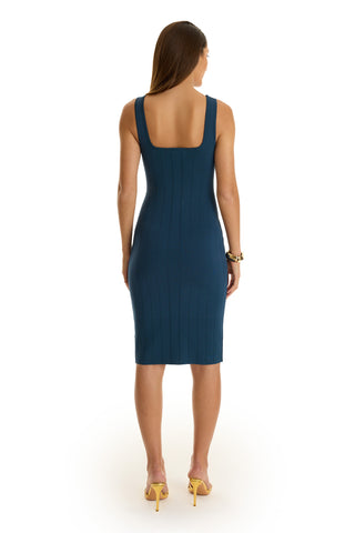 A woman with long brown hair wears a fitted, sleeveless Helena Button Dress - Ocean, featuring gold dome buttons and a square neckline. She pairs it with yellow heels and a bracelet, standing against a white background, facing away.