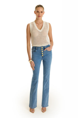 A woman faces forward wearing a sleeveless white crochet top, Aubrey Bootcut Jean - Medium Blue with gold buttons, nude heels, gold jewelry, hair pulled back, and one hand in her pocket.