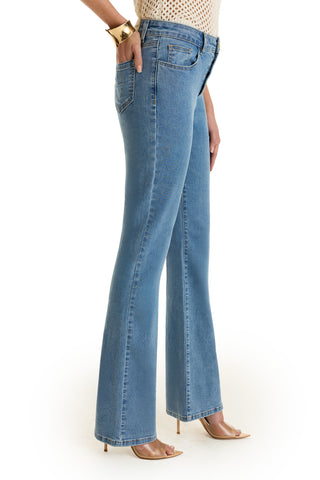 A woman poses in profile against a white background, wearing the Aubrey Bootcut Jean - Medium Blue, a cream crochet top, nude heels, and a chunky gold bracelet. Her left hand rests in her pocket.