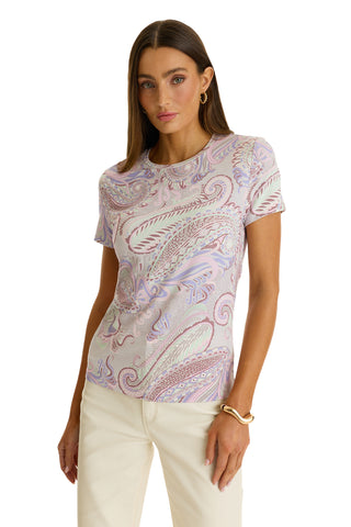 A woman with long brown hair wears the Amaya Ultra-Soft T-Shirt in Pastel Paisley and off-white pants, accessorized with gold hoop earrings and a gold bracelet, standing against a plain white background.