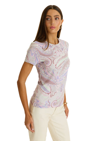 A woman with long brown hair models the Amaya Ultra-Soft T-Shirt in a pastel paisley pattern and light pants, standing sideways with one hand in her pocket, looking slightly left against a white background.