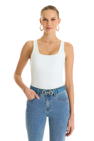 A woman with pulled-back hair wears the Loren Bodysuit in white, paired with blue jeans and a double-ring belt. She smiles softly with one hand in her pocket, standing against a plain white background and accessorized with large hoop earrings.