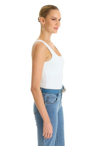 A woman stands in profile against a white background, her hair in a neat bun, wearing the Loren Bodysuit - White, blue high-waisted jeans with a belt, hoop earrings, and a necklace.