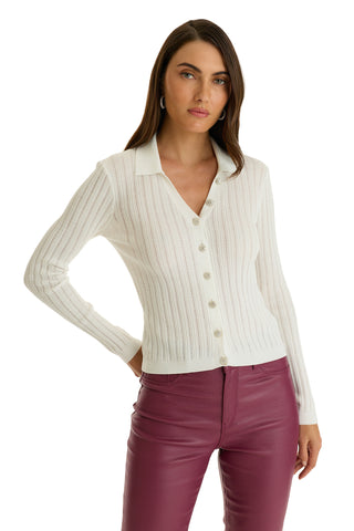 A woman confidently poses with one hand on her hip against a plain white background, wearing the Karolina Polo Shirt in white—featuring a ribbed knit, diamond buttons, and long sleeves—paired with high-waisted burgundy leather pants.