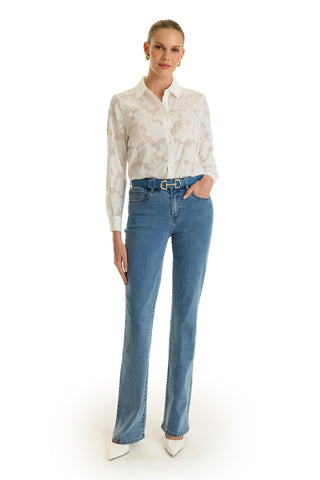 A woman stands against a white background, wearing the Stelle Button Up – White as a stylish layering piece, paired with blue high-waisted jeans, a belt, and white pointed-toe heels. Her hair is pulled back and her left hand rests in her pocket.