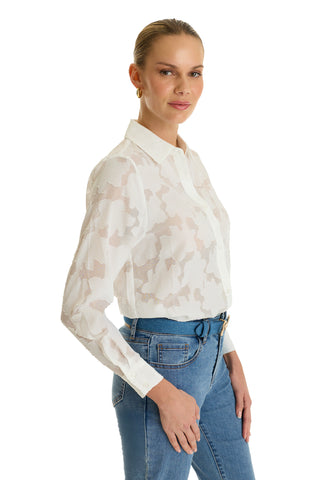 A woman with blonde hair tied back wears the Stelle Button Up - White, a sheer leaf-patterned blouse styled with blue jeans, standing in a side pose and smiling gently against a white background—the perfect layering piece.