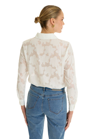 A woman with blonde hair in a low bun models the Stelle Button Up - White, a sheer patterned blouse ideal for layering, paired with light blue jeans. She faces away from the camera against a white background.