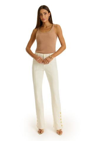 A woman with long hair and a neutral expression wears the Millie Knit Tank in mocha with a gold chain detail, high-waisted white pants with gold ankle buttons, and beige heeled sandals.