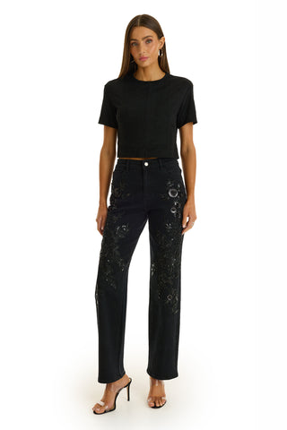 A woman with long brown hair wears a black short-sleeve top, the Stassi Floral Sequin Jean - Black, black heeled sandals, and hoop earrings, standing against a plain white background.