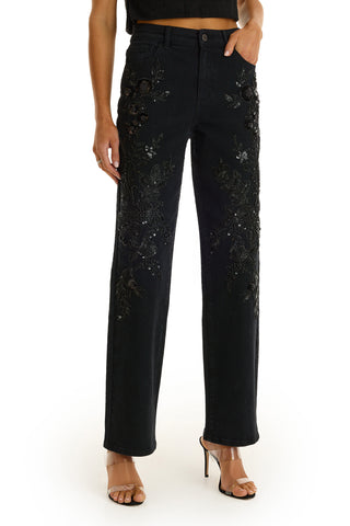 Wearing the Stassi Floral Sequin Jean in black, styled with a cropped black top and beige heels, a person stands against a white background.