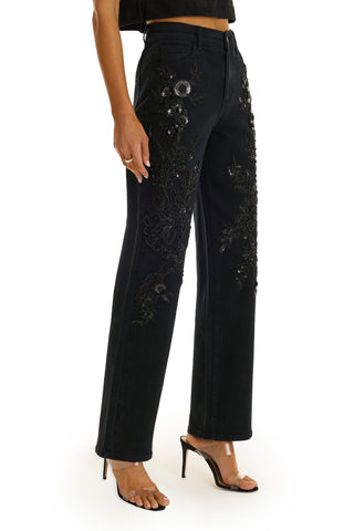 A woman models the Stassi Floral Sequin Jean in black—high-rise jeans with intricate black floral sequin embroidery—paired with a black crop top and heeled sandals against a white background.