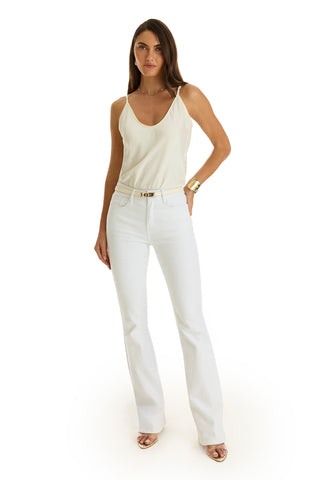 A woman faces forward, wearing a cream sleeveless top tucked into Aria Flare Jean - White with a belt. She has long brown hair, gold sandals, and gold jewelry, posing against a white background.